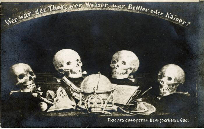 Black and white postcard with three human skulls sitting on a table with books and other items.